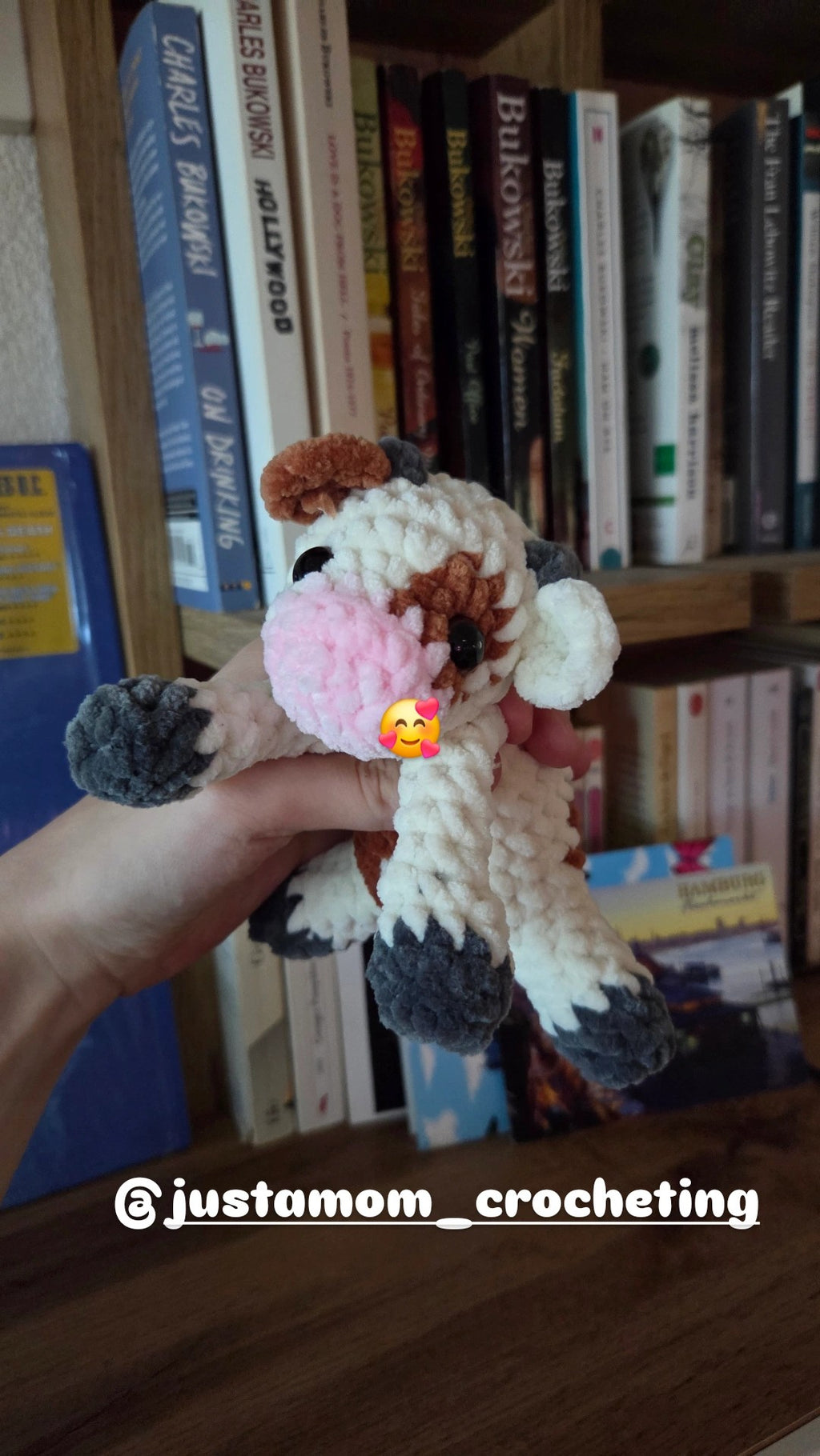 BrainRotPop handmade crochet cow plush toy in cream, brown, and pink chenille yarn held in front of a bookshelf.